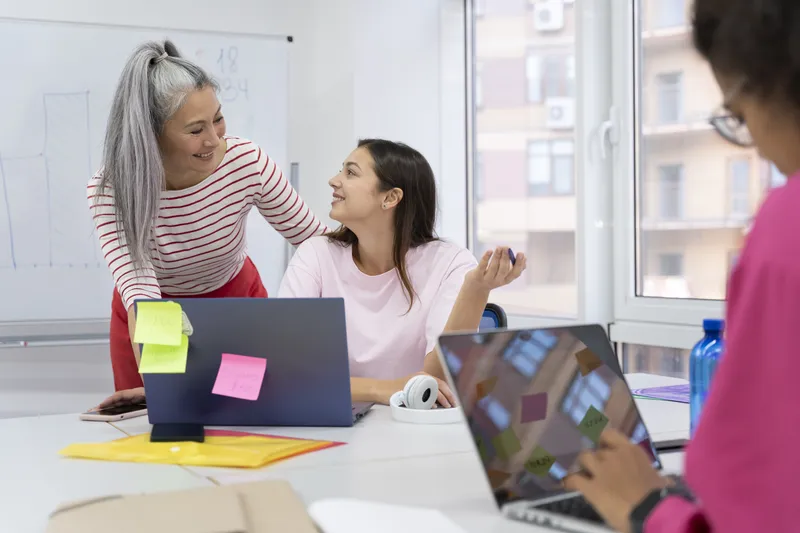 Two colleagues chat across a laptop at a bright office table