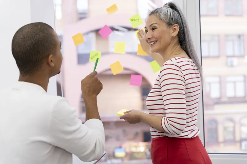 Two colleagues discuss colorful sticky notes on an office window
